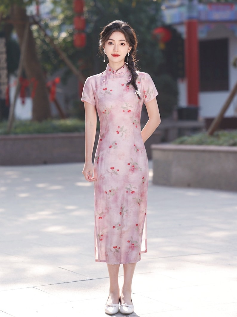 May include: A woman wearing a pink and white floral print cheongsam dress with a mandarin collar and short sleeves. She is standing on a light gray tiled surface.