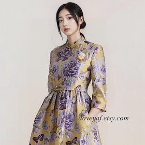 Peut inclure: Robe jaune à motifs floraux avec des roses violettes et des papillons. La robe a un col mandarin, des manches trois-quarts et une taille ajustée avec une petite ceinture. Une petite broche fleur est épinglée sur la poitrine. doveyaf.etsy.com est visible.