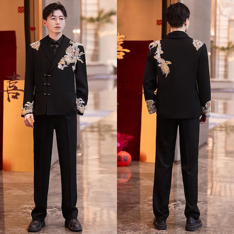 May include: Black suit with gold and white embroidery on the shoulders and sleeves. The suit includes a double-breasted jacket and matching pants. The jacket has a mandarin collar and decorative buttons. The suit is worn with a black shirt.