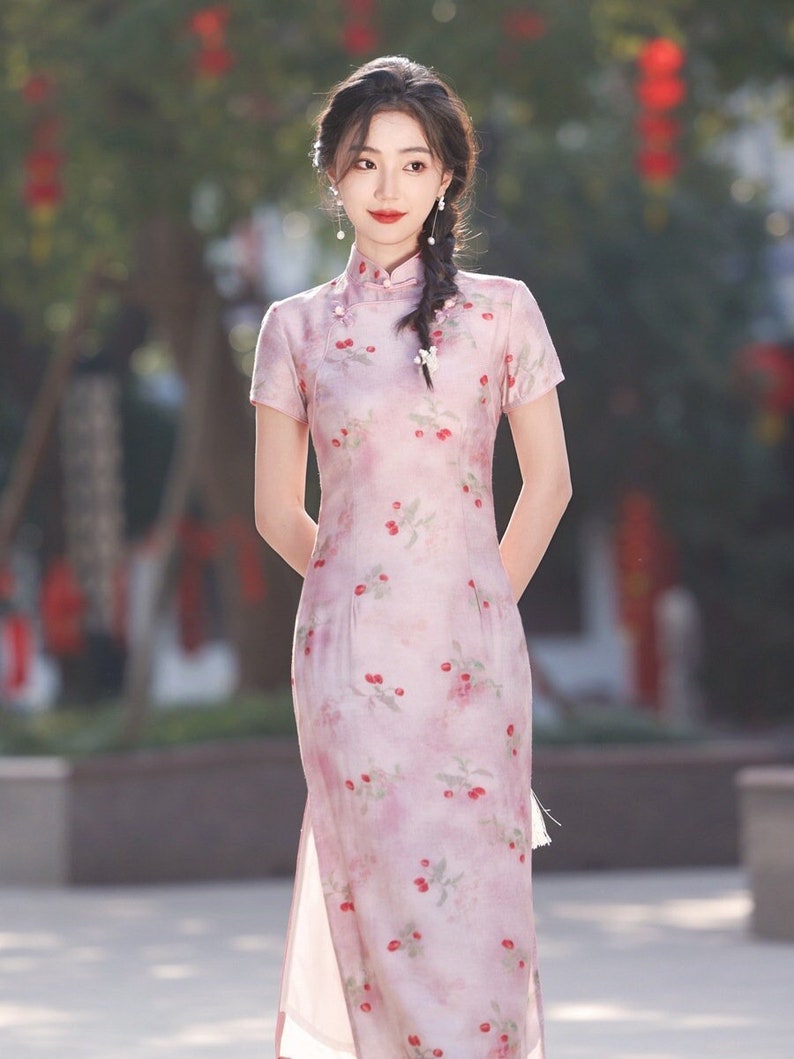 May include: A woman wearing a pink and white floral print cheongsam dress with a high collar and short sleeves. The dress has a side slit and a tassel detail. The woman has her hair pulled back in a braid and is wearing pearl earrings.