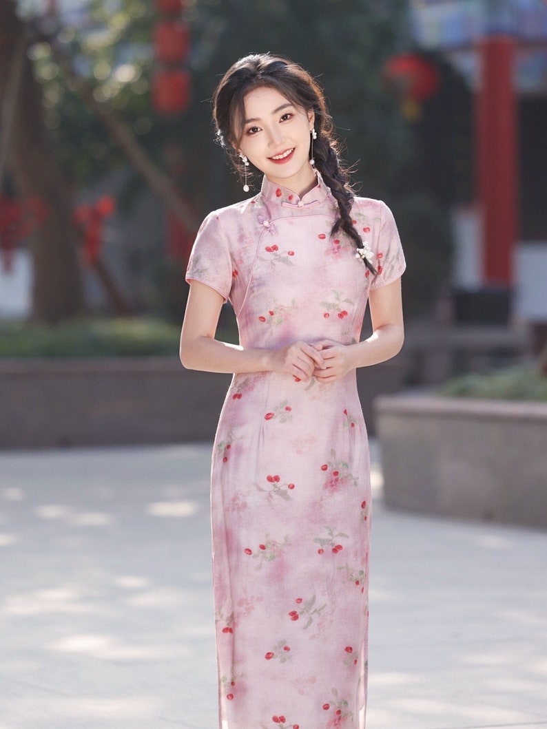 May include: A woman wearing a pink and white floral dress with a mandarin collar. The dress has a cherry print and is knee-length.