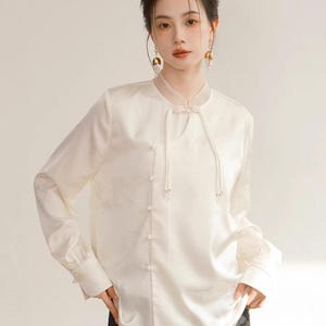 Könnte beinhalten: Eine cremefarbene, langärmlige Bluse mit Stehkragen und zarten perlenartigen Knöpfen. Die Bluse hat ein dezentes, geprägtes Muster und einen Kordelverschluss am Ausschnitt. Die Ärmel haben geknöpfte Manschetten, und der Gesamtstil ist elegant und raffiniert.