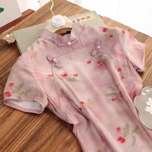 May include: A pink and white floral print dress with short sleeves and a mandarin collar. The dress has a button closure at the neck and is decorated with small, pink flower embellishments.