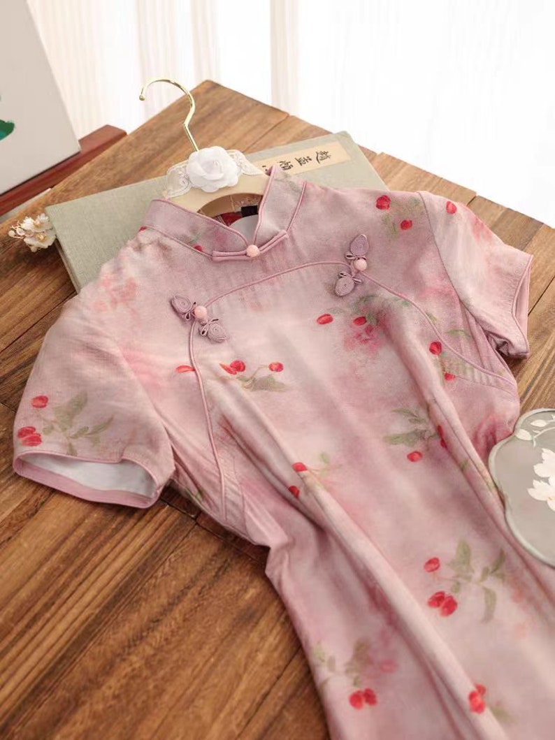May include: A pink and white floral print dress with short sleeves and a mandarin collar. The dress has a button closure at the neck and is decorated with small, pink flower embellishments.