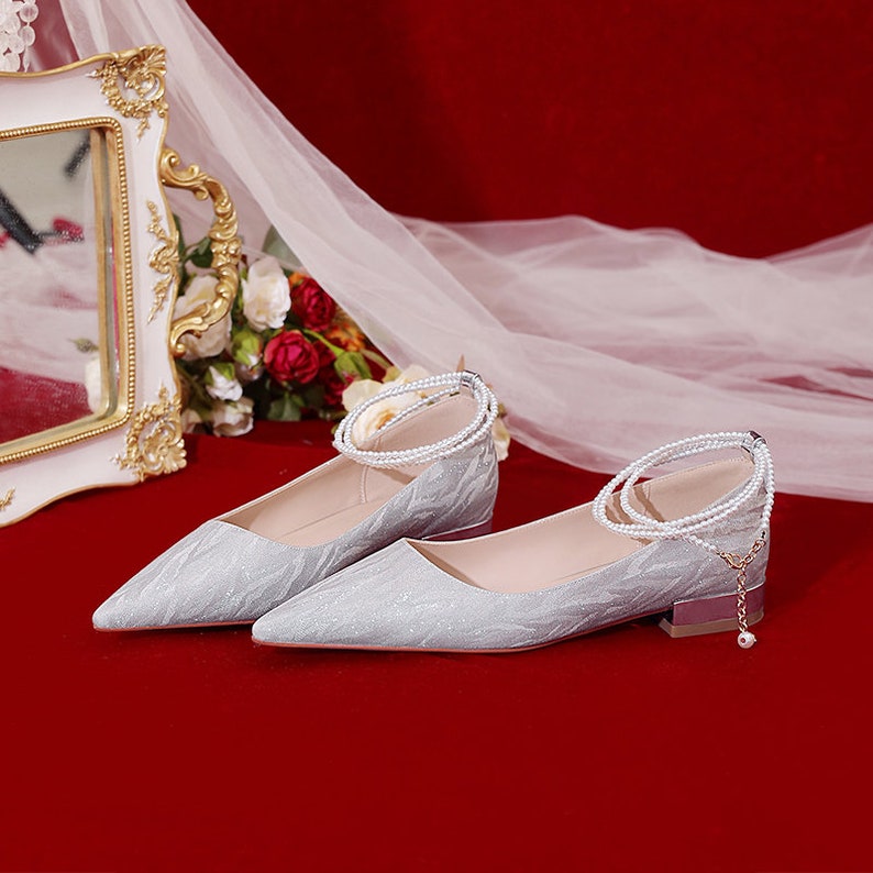 May include: A pair of silver glitter pointed-toe flats with a low heel. The shoes have a pearl ankle strap and a small pearl charm.