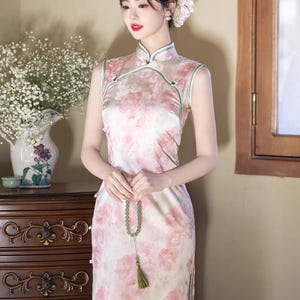 May include: A sleeveless, floral-print dress in shades of pink and white. The dress features a mandarin collar and a side slit. A woman is wearing the dress and holding a beaded necklace with a tassel. The background includes a vase of flowers and a wooden dresser.