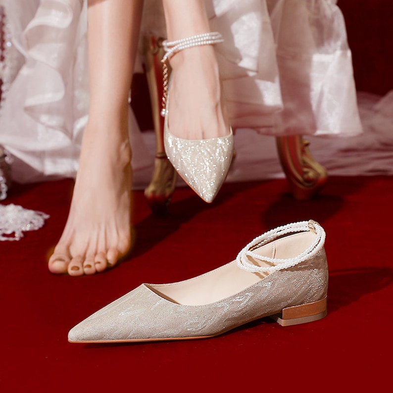 May include: A pair of silver sparkly pointed-toe flats with a pearl ankle strap. The shoes are on a red carpet.
