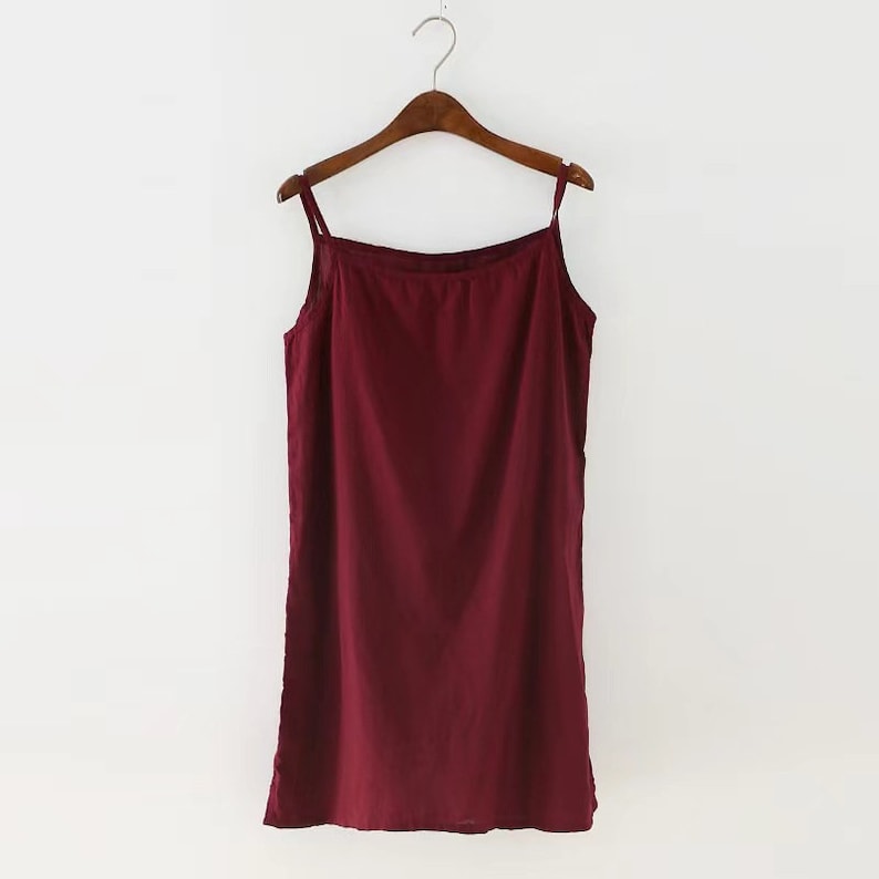 K&ouml;nnte beinhalten: Ein &auml;rmelloses Kleid in Burgunderrot mit einer schlichten, geraden Silhouette. Das Kleid h&auml;ngt an einem Holzb&uuml;gel.