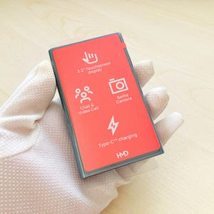 May include: A red rectangular device with a 3.2" touchscreen display, selfie camera, and Type-C charging port. The device also features chat and video call capabilities. The device is held in a gloved hand.