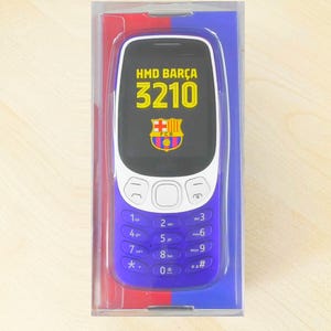 May include: A blue and white cell phone with the text "HMD BARÇA 3210" and a team logo on the screen. The phone is in a clear plastic case with red and blue accents. The phone has a keypad with blue buttons.