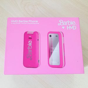May include: A pink box featuring a pink Barbie phone. The box has the text "HMD Barbie Phone" and "A real phone, without the distractions." The Barbie logo is visible. The phone is shown in two views, closed and open.