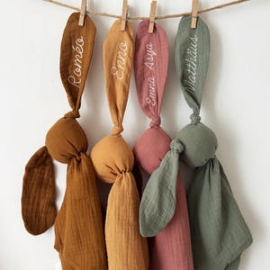 May include: Four knotted fabric bunny toys in shades of brown, gold, pink, and green hang from a clothesline. Each bunny has long, ear-shaped fabric with a name embroidered in white: Rom&eacute;o, Enno, Emma Asya, and Matthias.