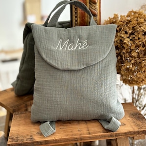 May include: A sage green muslin backpack featuring a top flap with the name Mah&eacute; embroidered in white thread. The bag has a soft, crinkled texture, a top handle, and two shoulder straps. A small white fabric tag is visible on the side of the bag.
