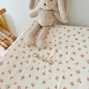 Peut inclure: Une couverture en mousseline de coton blanche avec un motif floral rose et le nom "Charlotte" brodé en fil doré. Un lapin en peluche beige est assis sur la couverture.