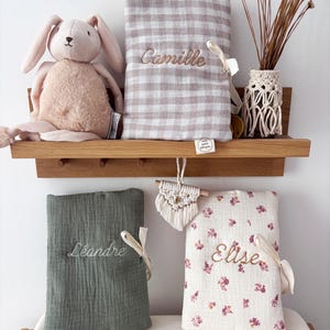 May include: A plush rabbit toy, a beige and white gingham fabric cover embroidered with the name Camille, a solid sage green fabric cover with the name Léandre, and a white fabric cover with a pink floral pattern and the name Elise.