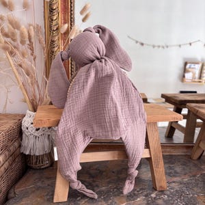 May include: A dusty rose-colored baby security blanket shaped like a bunny. The blanket is made of a soft, textured fabric with long, floppy ears and knotted feet. It is resting on a small wooden stool.