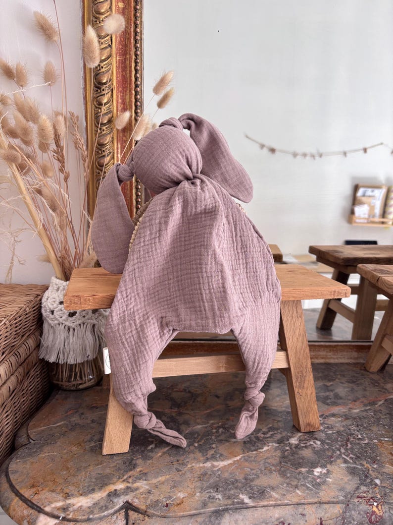 May include: A dusty rose-colored baby security blanket shaped like a bunny. The blanket is made of a soft, textured fabric with long, floppy ears and knotted feet. It is resting on a small wooden stool.
