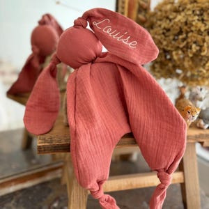 May include: A coral-colored bunny-shaped baby comforter with long, knotted ears and a soft, textured fabric. The name "Louise" is embroidered in white script on one ear. The comforter is displayed on a small wooden stool.