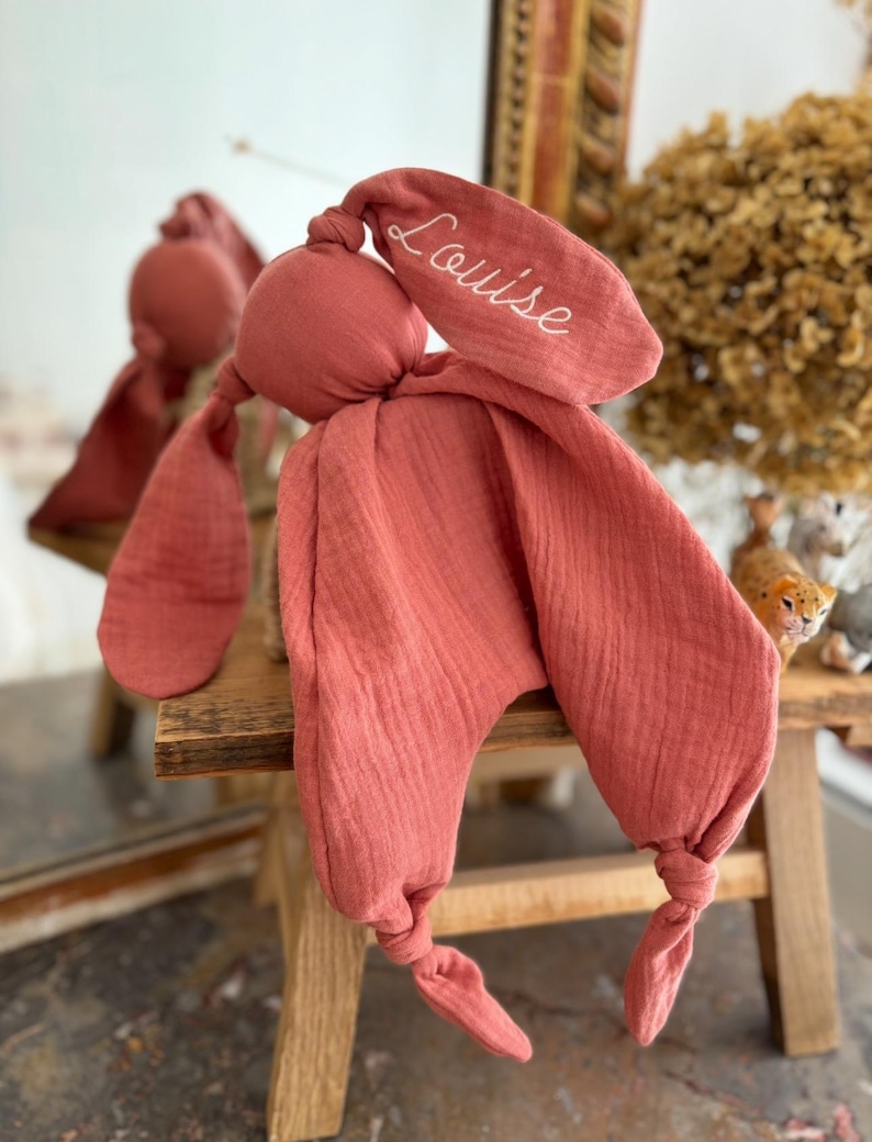 May include: A coral-colored bunny-shaped baby comforter with long, knotted ears and a soft, textured fabric. The name "Louise" is embroidered in white script on one ear. The comforter is displayed on a small wooden stool.
