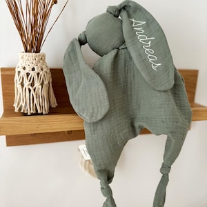 May include: Sage green baby security blanket in the shape of a bunny. The bunny has long ears, one of which is embroidered with the name "Andreas" in white. The blanket is made of a soft, textured fabric and has knotted ends.