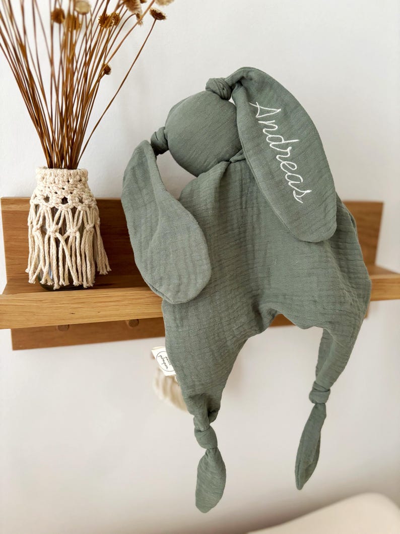 May include: Sage green baby security blanket in the shape of a bunny. The bunny has long ears, one of which is embroidered with the name "Andreas" in white. The blanket is made of a soft, textured fabric and has knotted ends.