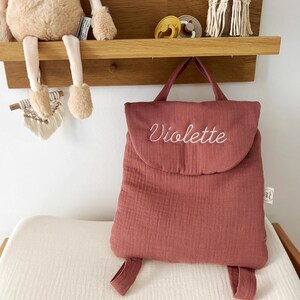 May include: A dusty rose-coloured fabric backpack featuring a top flap with the name Violette embroidered in white cursive script. The backpack has a top handle, two shoulder straps, and a small white fabric tag on the right side seam.