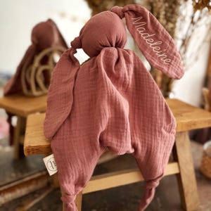 May include: A dusty rose-colored bunny-shaped baby comforter. The comforter has a knotted head and ears, with the name "Madeleine" embroidered on one ear. The fabric is textured and soft, with a tag that says "petit pompon".