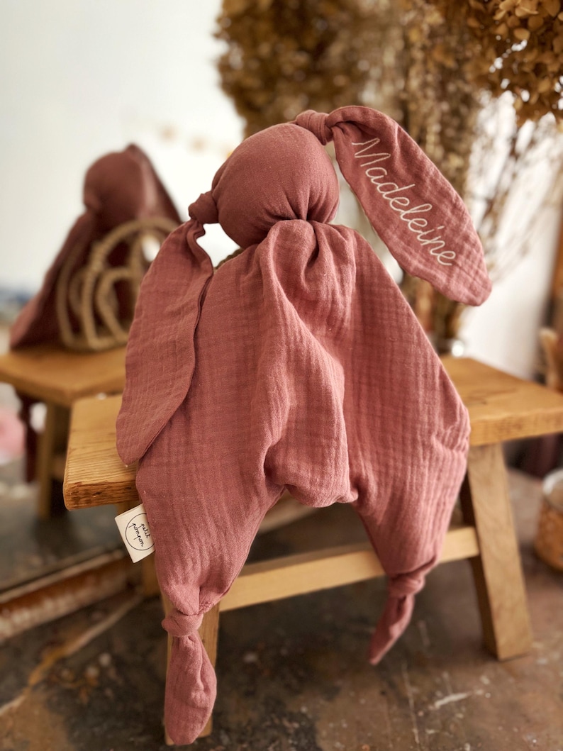 May include: A dusty rose-colored bunny-shaped baby comforter. The comforter has a knotted head and ears, with the name "Madeleine" embroidered on one ear. The fabric is textured and soft, with a tag that says "petit pompon".