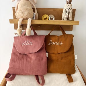 May include: Two soft fabric backpacks with top handles and shoulder straps. The left backpack is dusty rose with the name Alix embroidered in white. The right backpack is mustard yellow with the name Jonas embroidered in white. Both feature a flap.