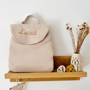 May include: A beige muslin backpack with a top handle and a rounded flap featuring the embroidered name Lucas in gold thread. The backpack is displayed alongside two round pacifiers and a small vase with dried flowers on a wooden shelf.