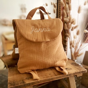 May include: A mustard yellow muslin backpack featuring a top flap with the name Janna embroidered in white cursive script. The bag includes a top handle, two shoulder straps, and a small white rectangular brand tag on the side seam.