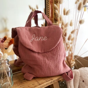 May include: A dusty rose-coloured fabric backpack featuring a rounded flap with the name Jane embroidered in white cursive script. The backpack has a top handle and adjustable shoulder straps, crafted from a soft, textured muslin-like material.
