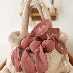 May include: A beige fabric tote bag with a matching set of bunny ear toys in a dusty rose color. The bunny ears are embroidered with the name "Mila". The bag has two handles and is sitting on a white surface.