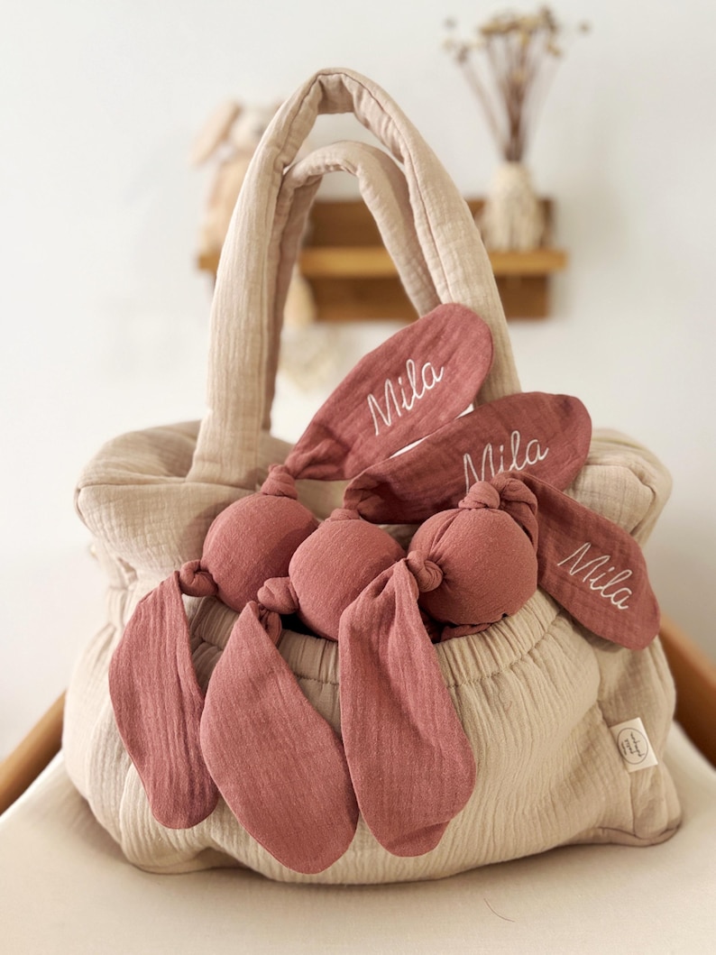 May include: A beige fabric tote bag with a matching set of bunny ear toys in a dusty rose color. The bunny ears are embroidered with the name "Mila". The bag has two handles and is sitting on a white surface.