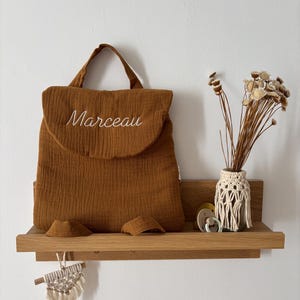 May include: A rust-coloured fabric backpack with a top handle and a flap closure featuring the embroidered name Marceau in white script. The backpack is displayed on a wooden shelf alongside a small macrame-wrapped vase with dried flowers.