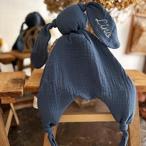 May include: A navy blue bunny-shaped baby comforter with long floppy ears. The comforter is made of a soft, textured fabric. The name "Lina" is embroidered in white on one ear. The comforter is sitting on a small wooden stool.