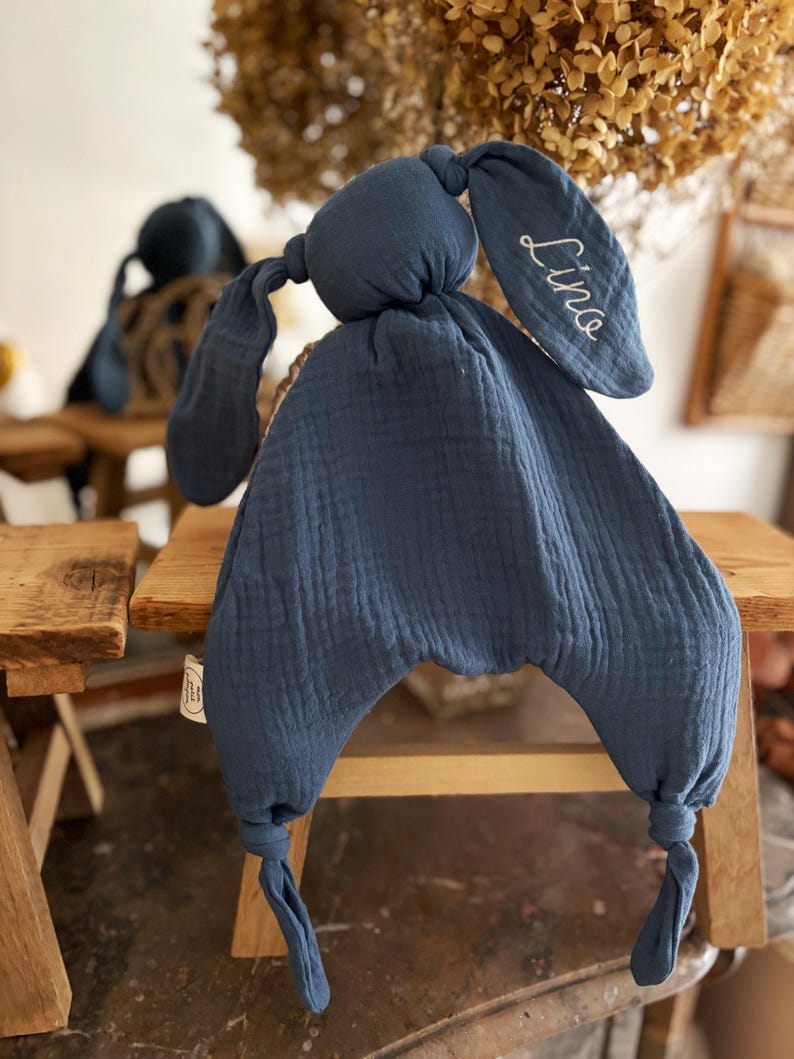 May include: A navy blue bunny-shaped baby comforter with long floppy ears. The comforter is made of a soft, textured fabric. The name "Lina" is embroidered in white on one ear. The comforter is sitting on a small wooden stool.