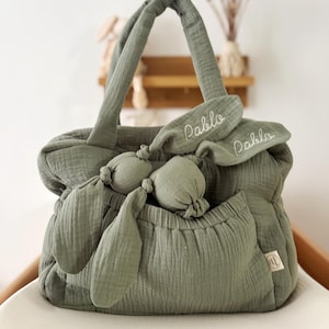 May include: Sage green baby bag with matching bunny ears and a small pocket. The bag has two handles and a soft, textured appearance. The name "Pablo" is embroidered on the bunny ears. A small tag is visible on the side of the bag.