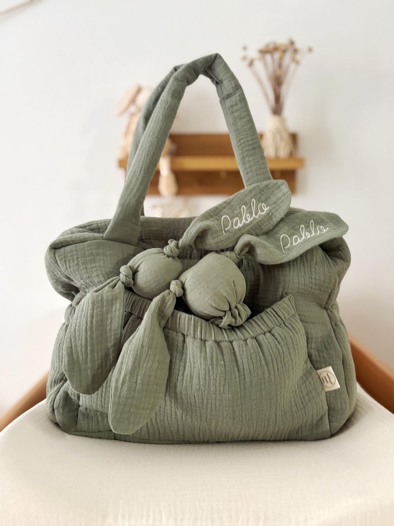 May include: Sage green baby bag with matching bunny ears and a small pocket. The bag has two handles and a soft, textured appearance. The name "Pablo" is embroidered on the bunny ears. A small tag is visible on the side of the bag.