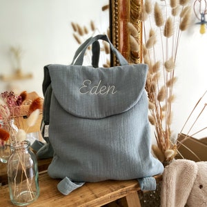 May include: A dusty blue fabric backpack featuring a top flap with the name Eden embroidered in white script. The backpack has two top handles and adjustable shoulder straps, constructed from a soft, textured material with a simple fold-over closure.