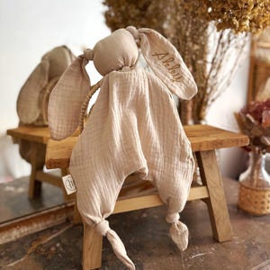 May include: A beige bunny-shaped baby comforter with long, floppy ears. The ear has the name "Alelly" embroidered in gold thread. The comforter is made of soft, textured fabric and has knotted ends. A small tag is attached.