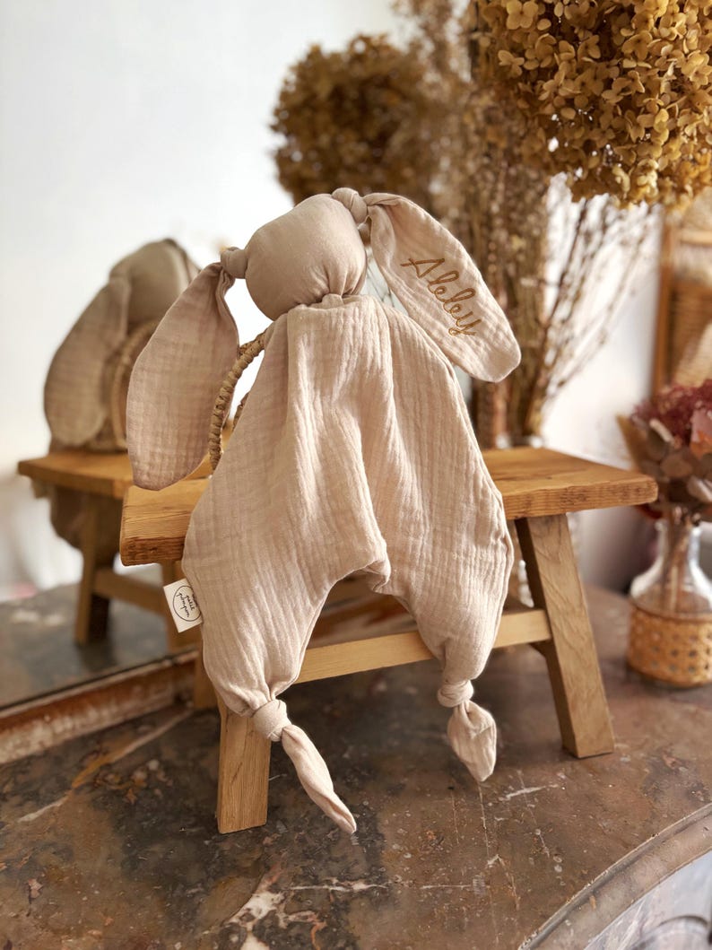 May include: A beige bunny-shaped baby comforter with long, floppy ears. The ear has the name "Alelly" embroidered in gold thread. The comforter is made of soft, textured fabric and has knotted ends. A small tag is attached.