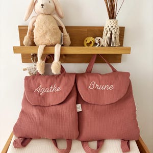 May include: Two dusty rose-coloured fabric backpacks with top handles and shoulder straps. Each backpack features a flap closure with the embroidered names Agathe and Brune in white cursive script. The bags are displayed below a wooden shelf.