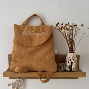 May include: A mustard yellow muslin backpack featuring a rounded flap with the embroidered name Balthazar in white thread. The bag has a top handle and two shoulder straps. Beside it sits a small vase with dried flowers and two pacifiers.