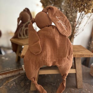 May include: A soft, brown bunny-shaped baby comforter. The comforter has long, floppy ears, one of which is embroidered with the name "Lyam" in white script. The body is a large, textured square with knotted corners, designed for comfort and security.