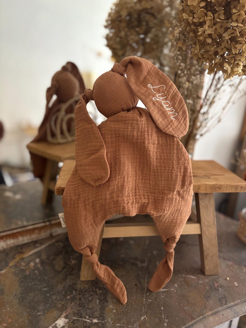 May include: A soft, brown bunny-shaped baby comforter. The comforter has long, floppy ears, one of which is embroidered with the name "Lyam" in white script. The body is a large, textured square with knotted corners, designed for comfort and security.