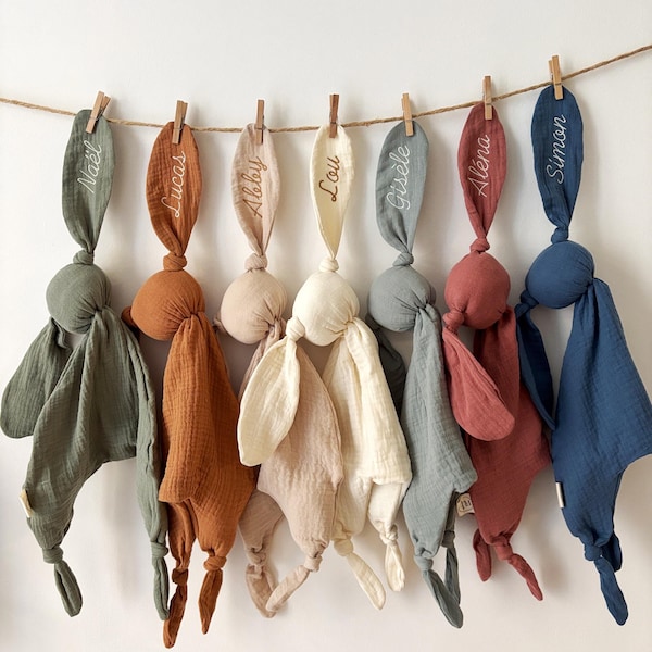 May include: Seven muslin security blankets with knotted corners and long ears, each embroidered with a name: Naël, Lucas, Abby, Lou, Gisèle, Aléna, and Simon. The blankets are in shades of olive, rust, beige, cream, sage, terracotta, and navy blue.