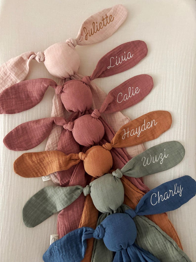 May include: A collection of soft, plush bunny toys in various colors, each with a name embroidered on one ear. The bunnies are in shades of pink, orange, green, and blue. The names include Juliette, Livia, Calie, Hayden, Wuzi, and Charly.