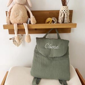 May include: A sage green fabric backpack featuring a top flap with the embroidered name C&ocirc;me in white script. The backpack has two shoulder straps and a top handle. It is displayed alongside a plush beige rabbit toy and two pacifiers.