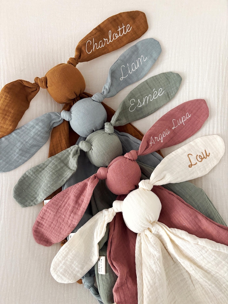 May include: A collection of personalized bunny-shaped baby toys in various colors, including brown, blue, green, pink, and white. Each bunny ear is embroidered with a name: Charlotte, Liam, Esmee, Argea Lupa, and Lou. The toys are made of soft, textured fabric.